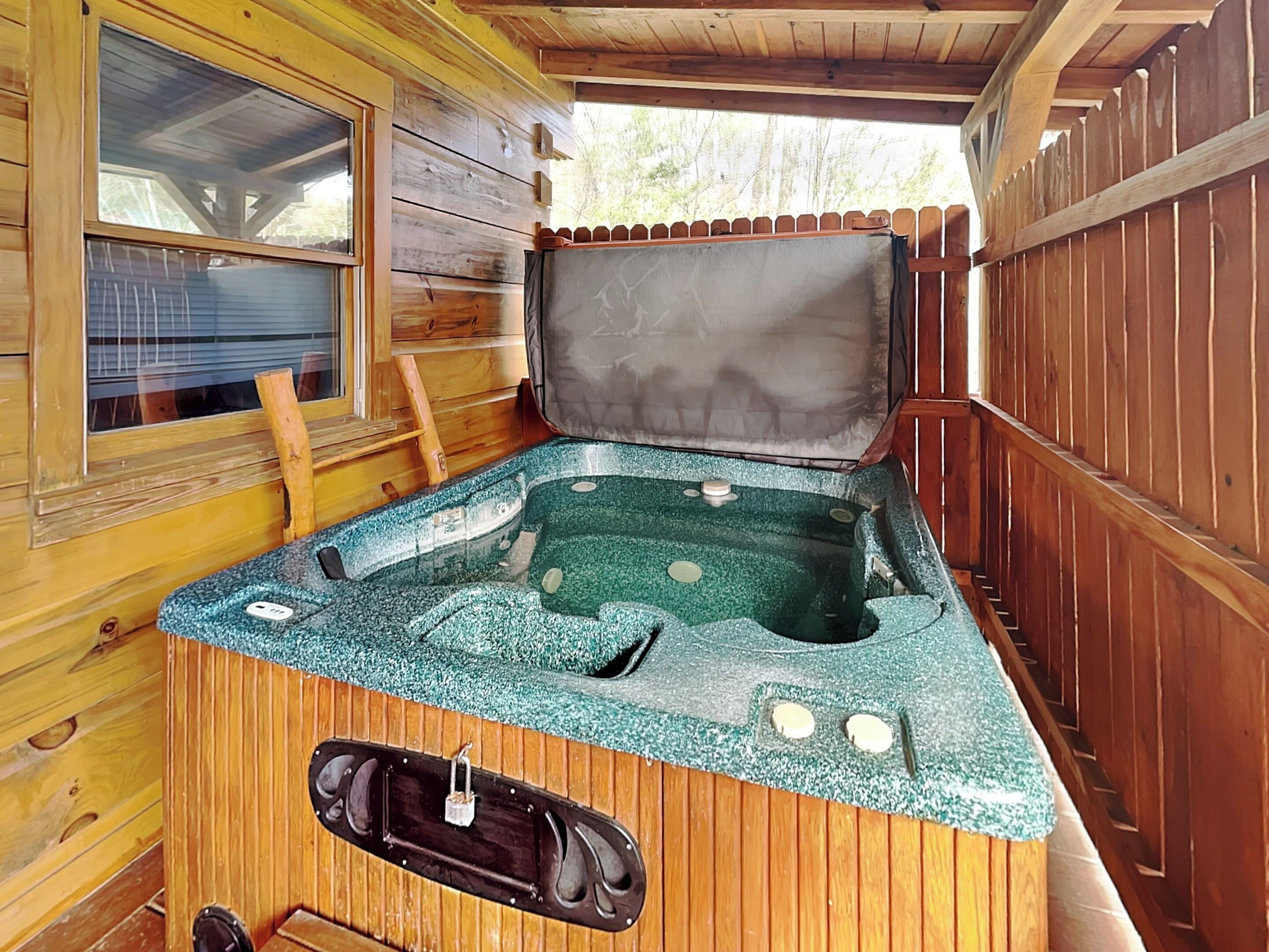 Une cabane en bois avec un jacuzzi au centre, une fenêtre sur le côté gauche et une terrasse en bois à l’extérieur.