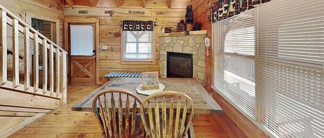 A wooden cabin interior with a stone fireplace, two rocking chairs, a dining table, and a window with blinds.