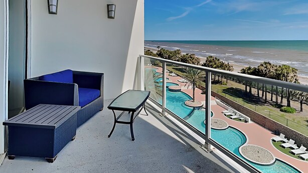 Un balcón con un sofá de mimbre y una pequeña mesa, con vistas a una zona de piscina con sillones y al océano.