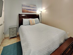A bedroom with a bed, a nightstand, a dresser, a painting on the wall, and a lamp. - Western Waves by Ryson Vacation Rentals (Galveston)
