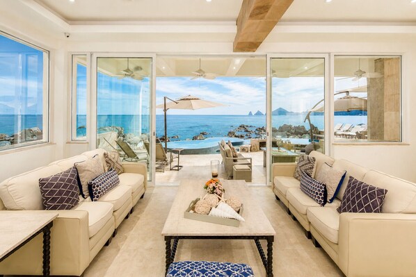 A spacious living area with a view of the ocean, two sofas, a coffee table, and a pool area visible through the glass doors.