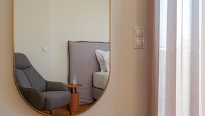A room with a large mirror, a grey armchair, a small table with a bottle, and a bed with white bedding.