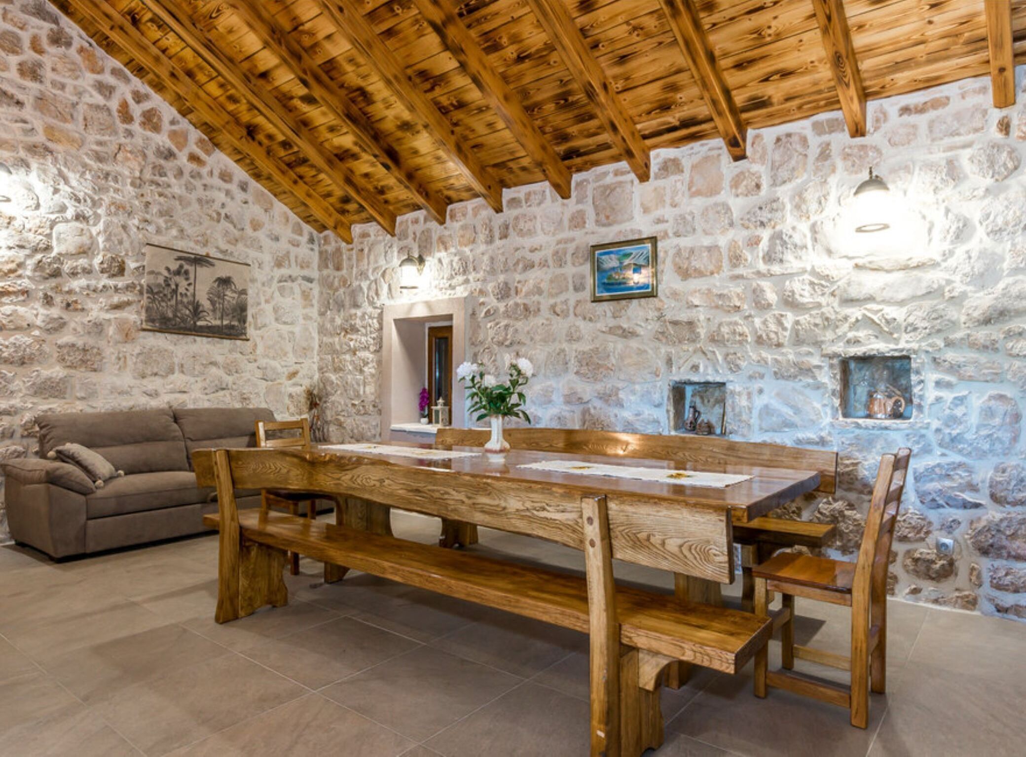 A rustic interior with a stone wall, wooden ceiling beams, a wooden dining table with benches, a sofa, and a small fireplace.