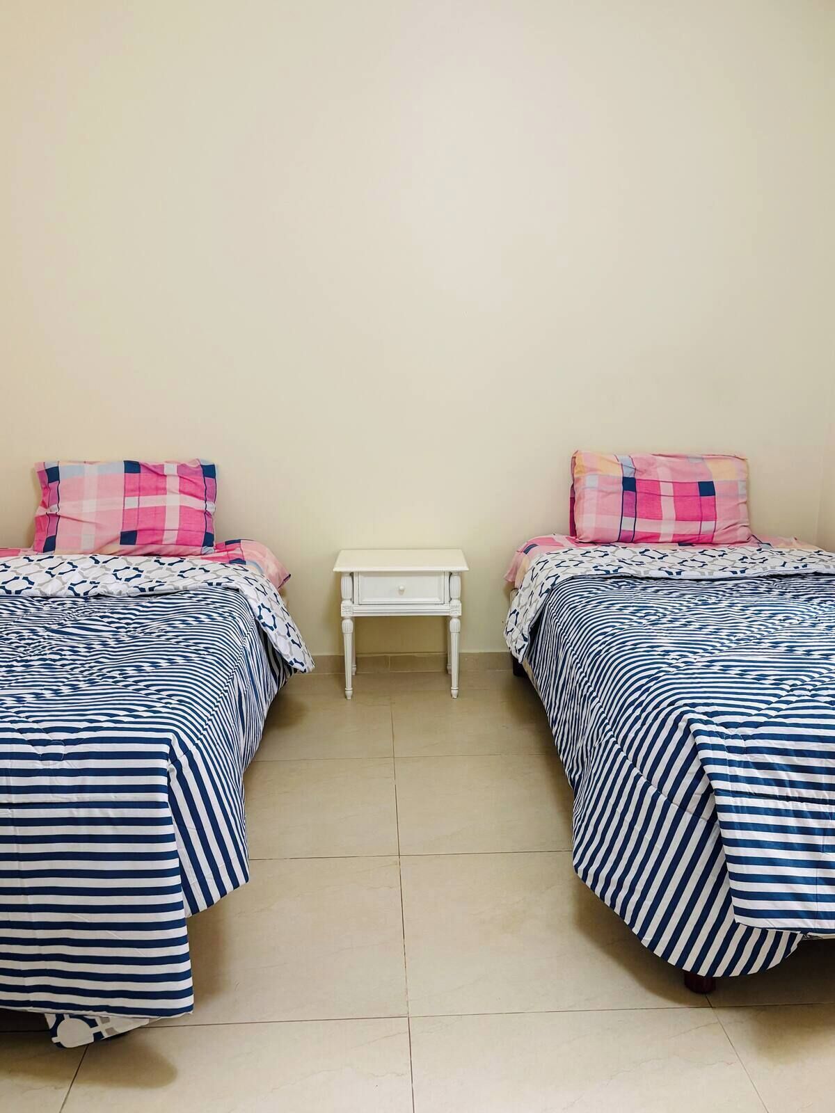 Een kamer met twee bedden, elk met gestreept beddengoed en roze kussens, een klein wit nachtkastje en een effen muur.