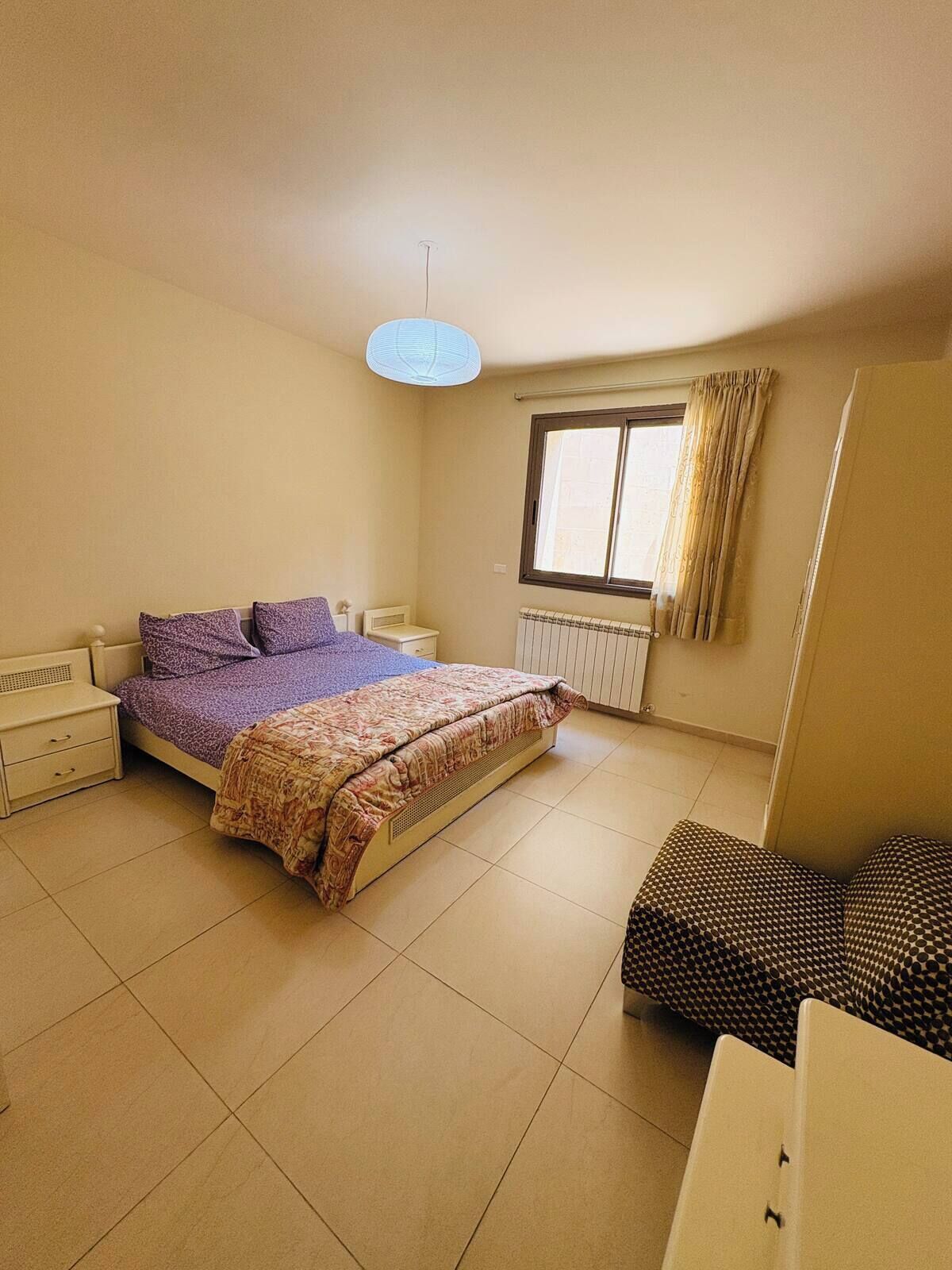Un dormitorio con cama, mesita de noche, radiador, ventana con cortinas y lámpara colgante.