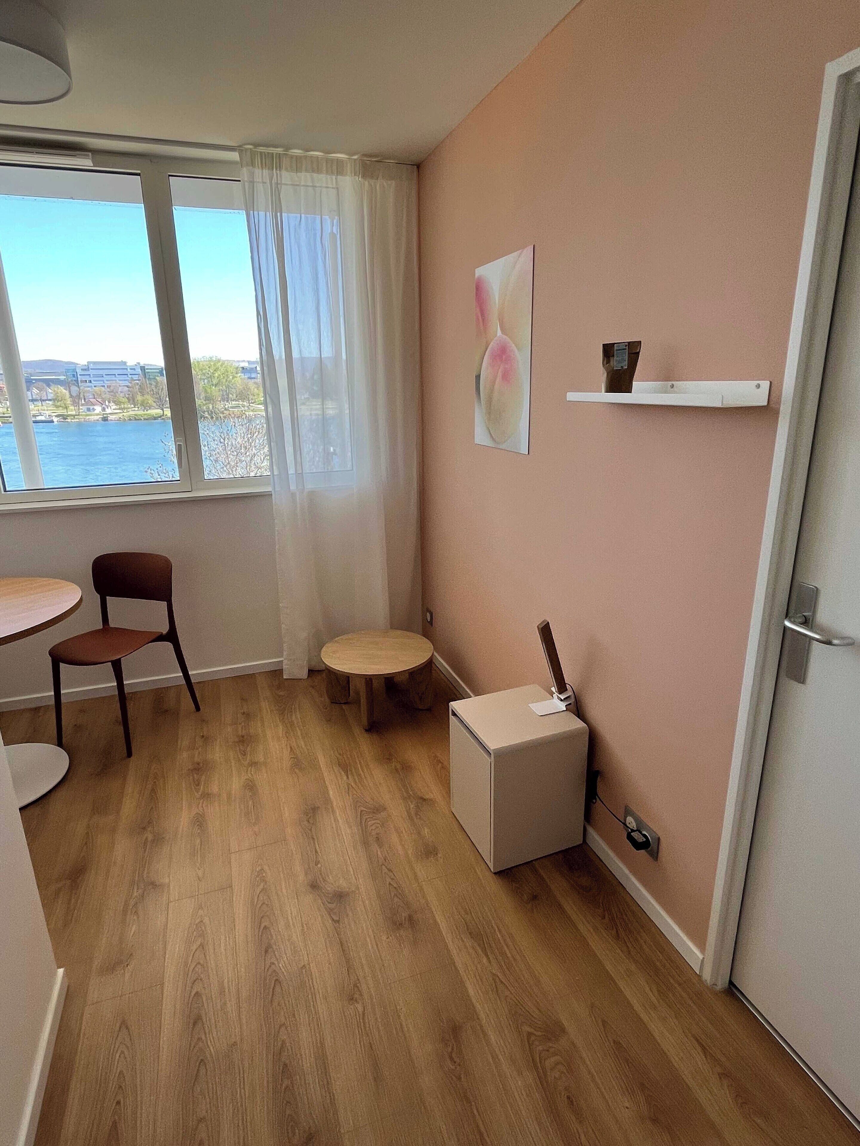 Une pièce avec un plancher en bois, une table ronde avec une chaise, un petit tabouret, une armoire blanche, une fenêtre donnant sur un plan d’eau et une étagère murale avec une tasse et un petit objet.