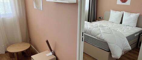 A bedroom with a bed, a bedside table, a small stool, a wall-mounted shelf, and a mirror.