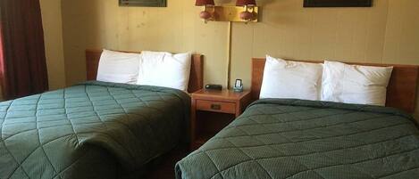 Habitación de hotel con dos camas individuales, cada una con una colcha verde acolchada y almohadas blancas. Entre las camas hay una mesita de noche de madera con una lámpara y un pequeño reloj. Las paredes están decoradas con cuadros enmarcados de paisajes.