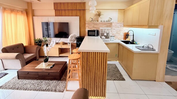Una sala de estar moderna con un sofá, una mesa de centro y un televisor. Se ve un área de cocina con un microondas, un lavabo y armarios de madera.