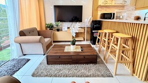 Una sala de estar moderna con sofá, una mesa de centro y televisor. Hay una zona de cocina con microondas y refrigerador.