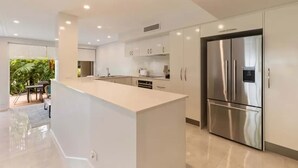 A modern kitchen with a central island, stainless steel appliances, and a view of a patio with greenery. - Port Douglas Mirage Villa 448 (Port Douglas)