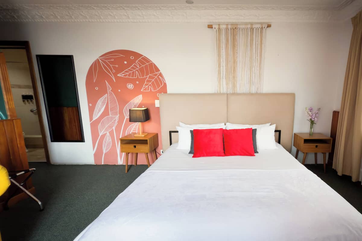 Habitación de hotel con una cama grande, una mesita de noche con lámpara, una silla de madera y una pared decorativa con motivos de hojas.