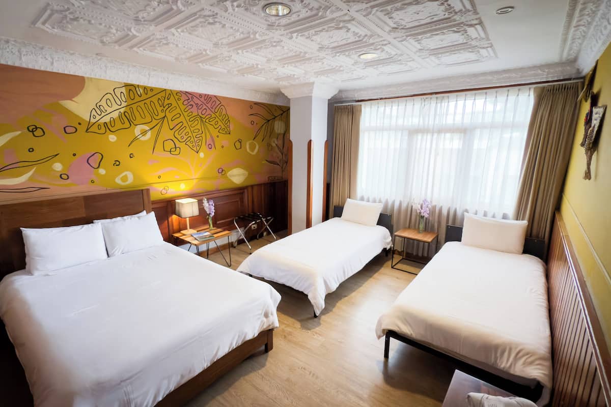Habitación de hotel con dos camas, un mural en la pared, una mesita con lámpara y una ventana con cortinas.