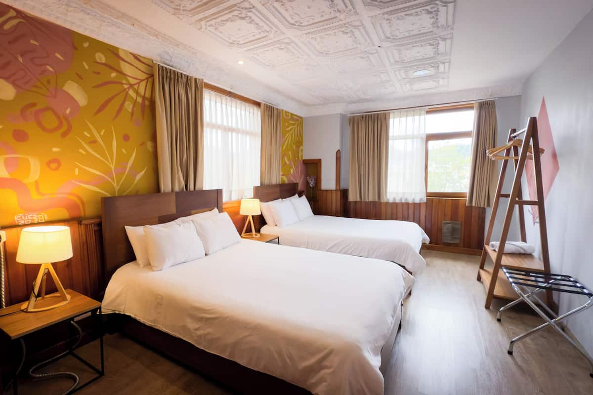 Habitación de hotel con dos camas, piso de madera, techo con diseños ornamentales y una ventana con cortinas.
