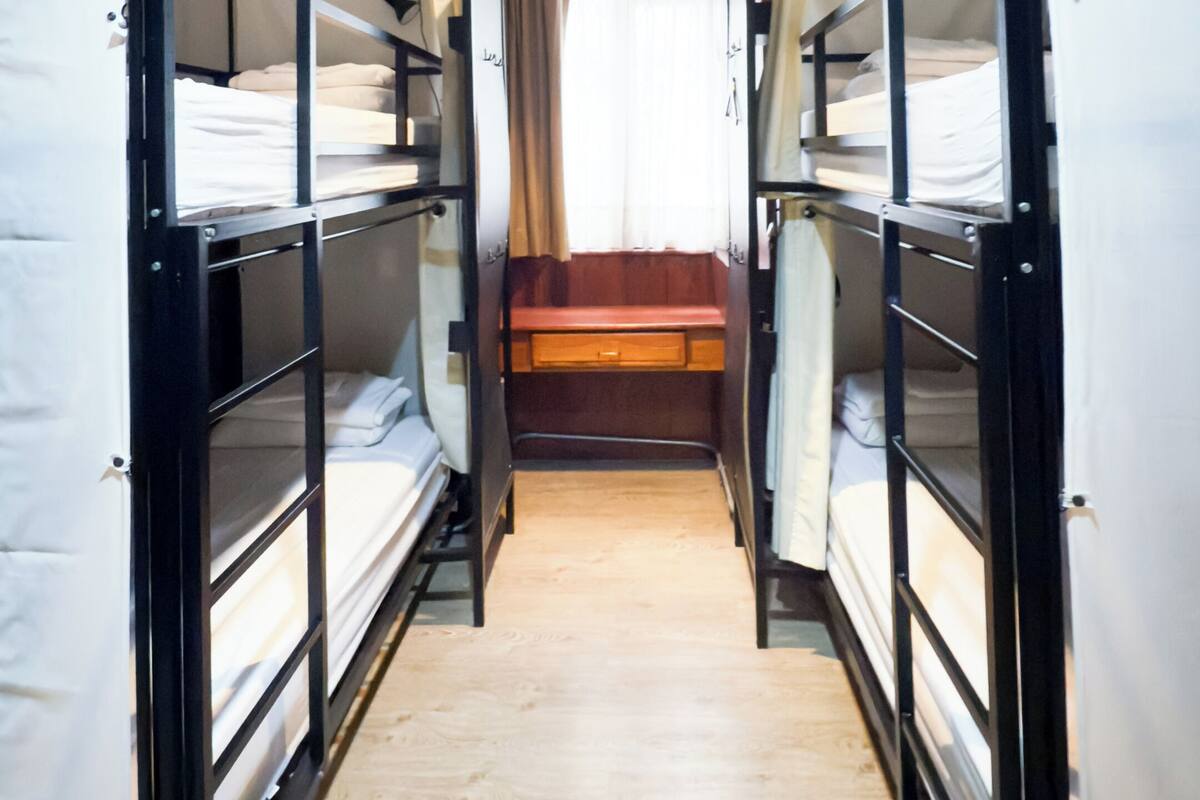 Un pasillo estrecho con literas a ambos lados, suelo de madera y una ventana con cortinas.