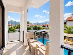 A balcony with a pool, wicker chairs, and a view of houses and mountains. - Bova Rooms (Kemer)