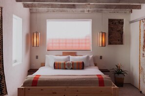 A bedroom with a wooden bed, white bedding, a large window with blinds, two wall-mounted lamps, and a potted plant.