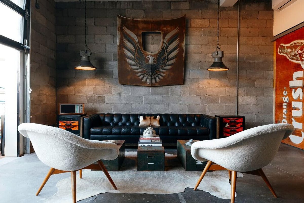 A modern lounge area with a black leather sofa, two white armchairs, and a coffee table. There is a large wall-mounted artwork above the sofa and a vintage orange sign on the right.