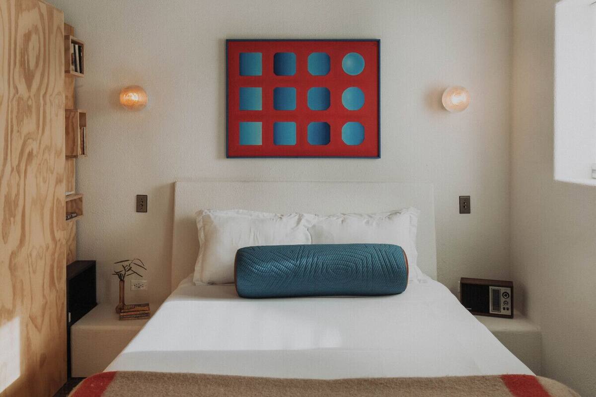 A bedroom with a bed, a blue pillow, a red and blue artwork, and a small bedside table with a lamp.