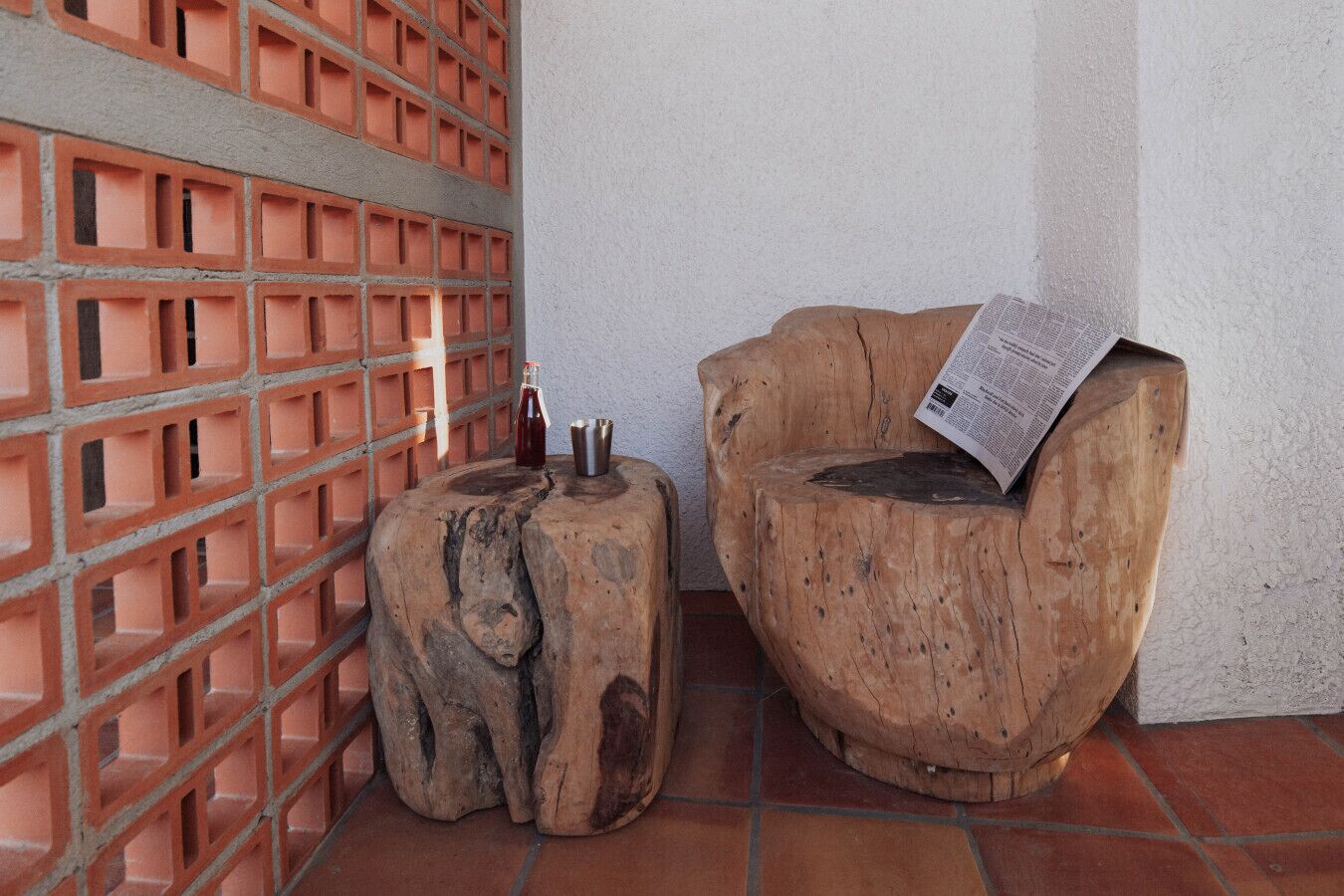 A corner with a brick wall, a wooden stool, and a wooden chair with a newspaper on it.