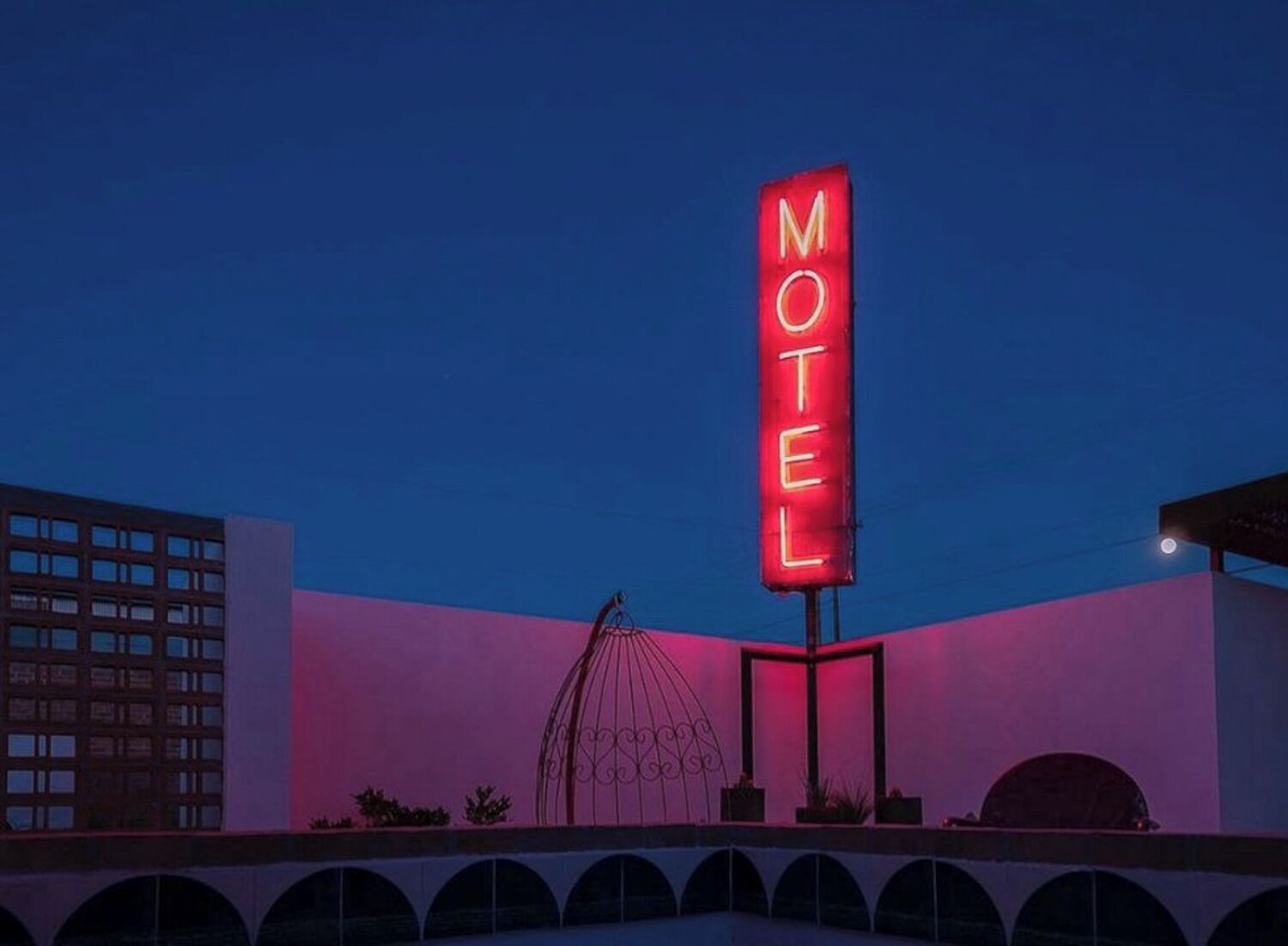 Ein Schild eines Motels mit Neonbeleuchtung, einer Metallskulptur und einem Gebäude mit rosa und violettem Farbton.