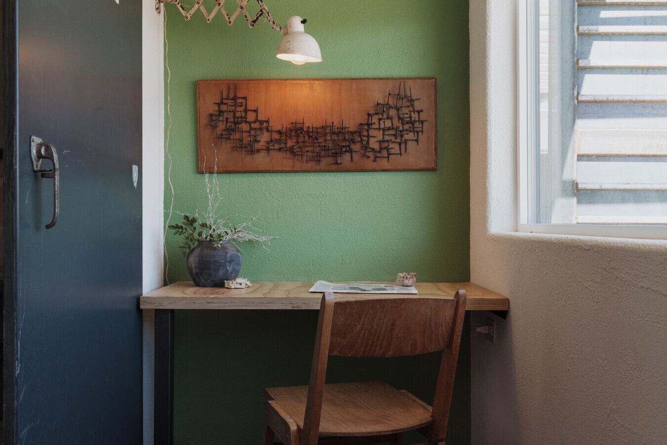 A corner room with a wooden desk, a chair, a potted plant, a wall-mounted abstract art piece, and a hanging lamp.