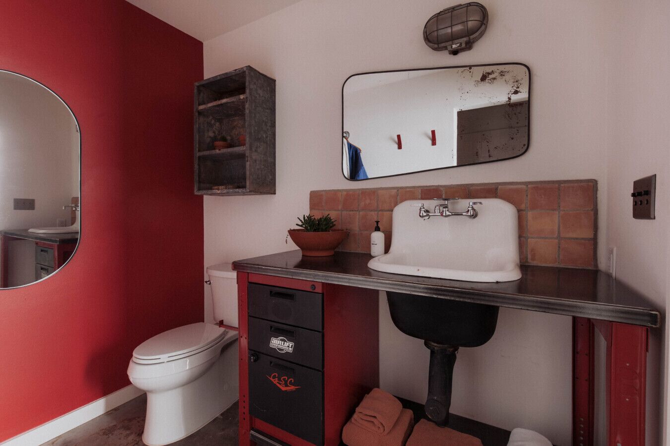 Un baño con inodoro blanco, un mueble rojo, un lavabo con una cubeta negra, un espejo y una planta en maceta.