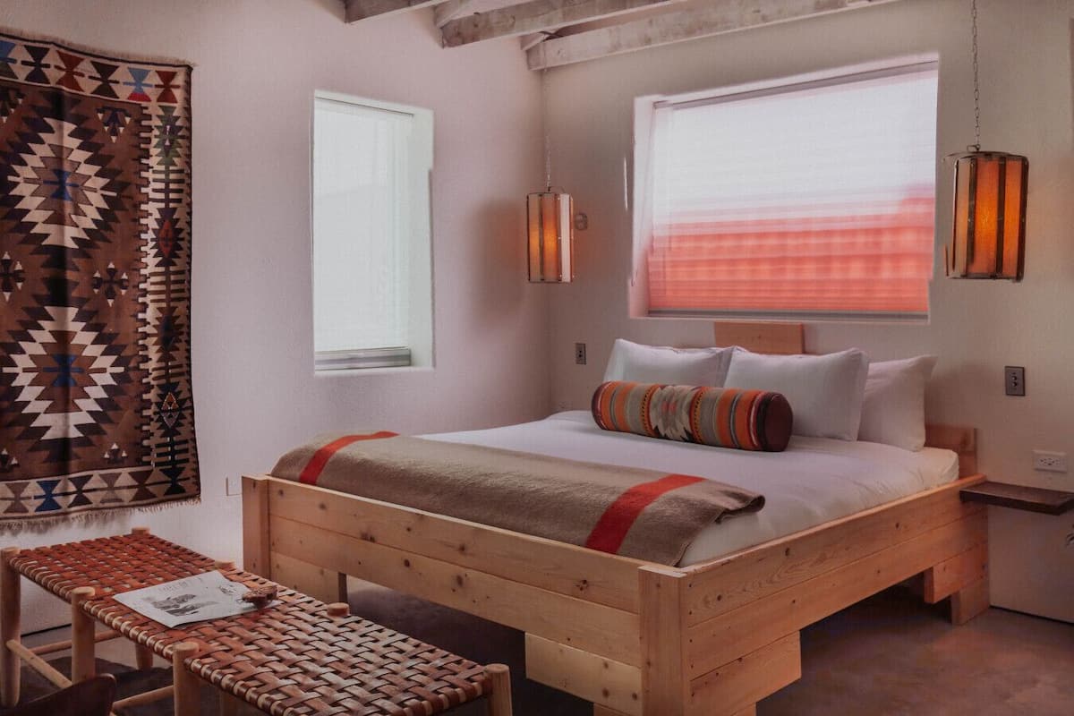 A cozy bedroom with a wooden bed, a woven bench, a patterned blanket, and a window with blinds.
