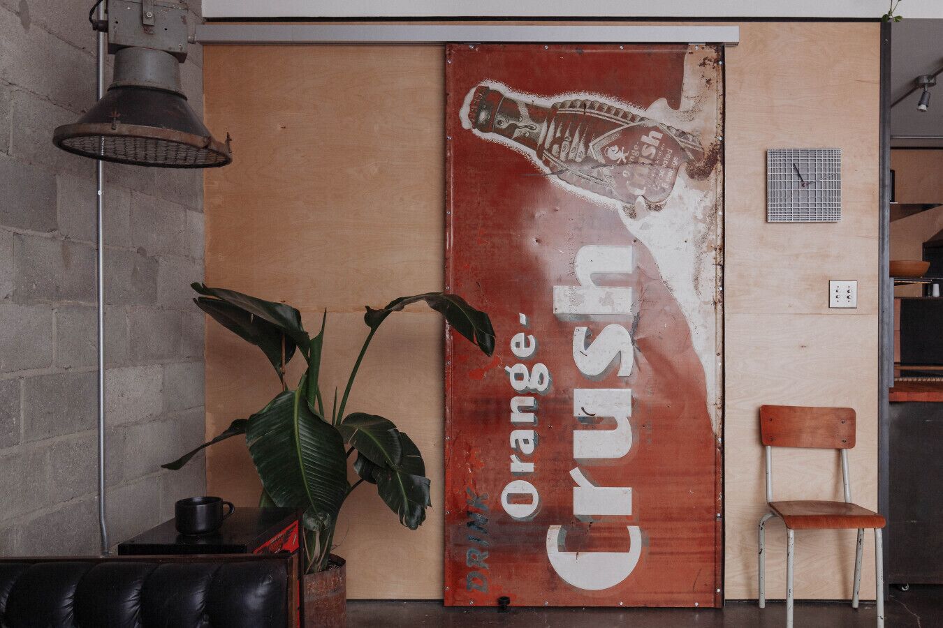 A wall with a large orange Crush beer advertisement, a potted plant, a metal chair, and a hanging light fixture.