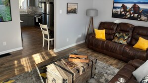 Ein modernes Wohnzimmer mit braunem Ledersofa, einem Essbereich mit weißem Stuhl, einer Küche mit grauen Schränken und einem großen Fenster.