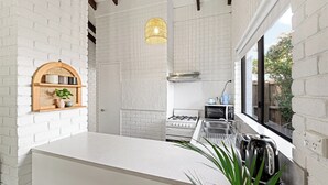 Une cuisine compacte aux murs en briques blanches, une étagère en bois ornée d’objets décoratifs, une cuisinière à gaz, un four encastré et une fenêtre donnant sur un paysage verdoyant.