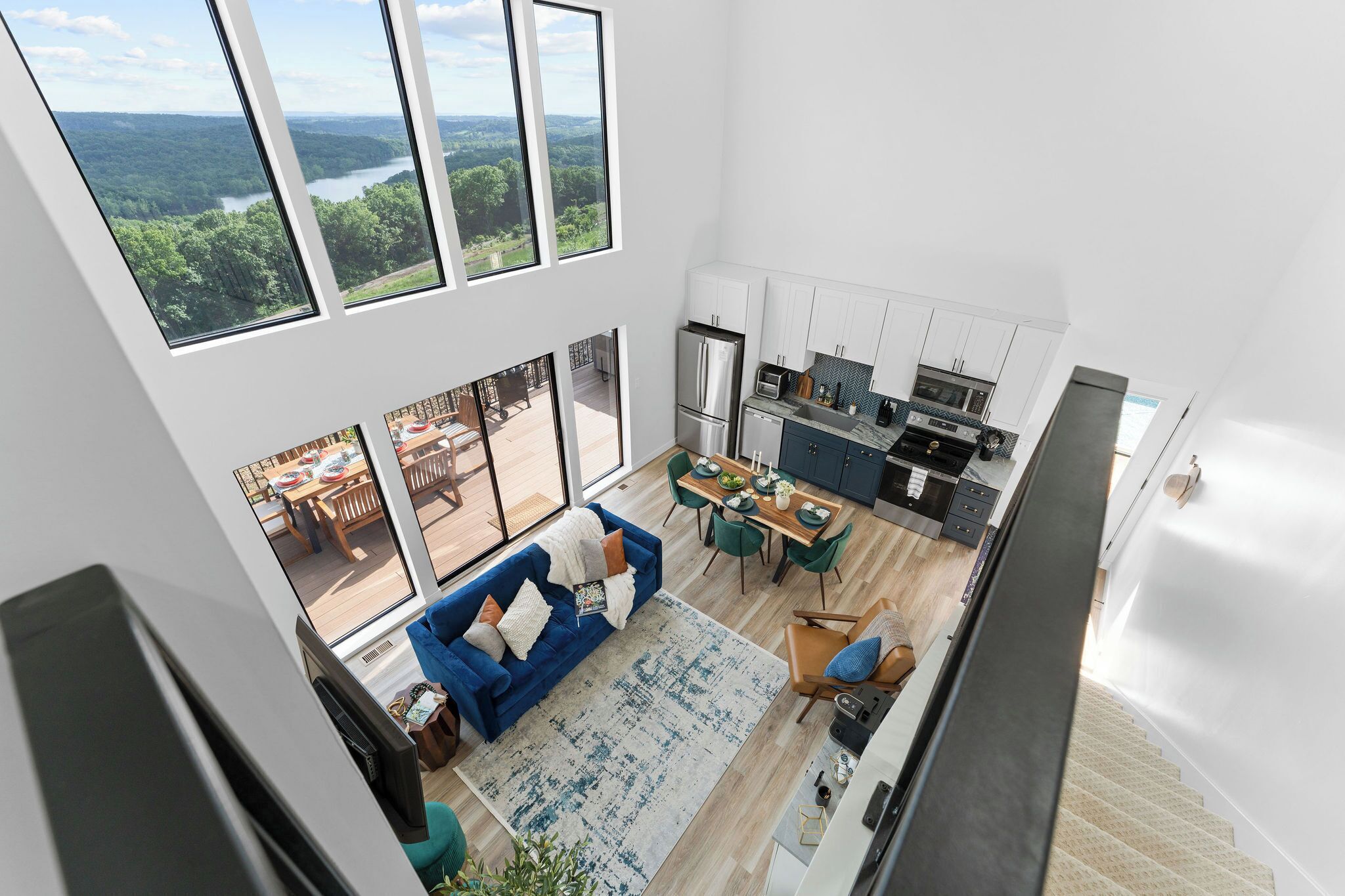A spacious living area with a large window overlooking a forest and river, a modern kitchen, and a dining area with green chairs.