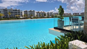 A clear blue pool with a glass edge, surrounded by lush greenery and a backdrop of modern resort buildings.