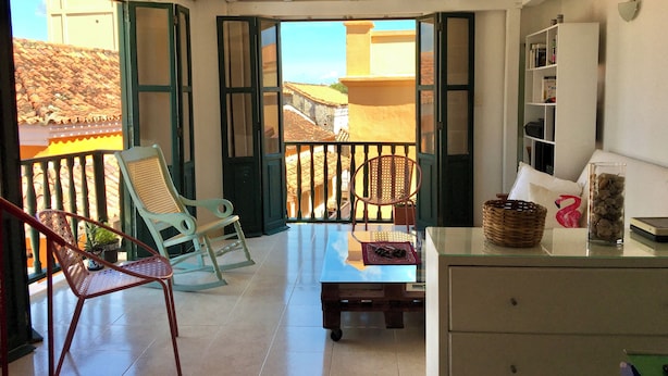 Een balkon met uitzicht op gebouwen, een tafel met tafelonderzetter, een stoel, een rieten mand en een glazen container op een kast.