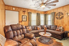 A room with a leather armchair, a sectional sofa, a round coffee table, and a ceiling fan. - The Lazy Spur | Cozy 2BR Cabin in Cowboy Capital (Bandera)