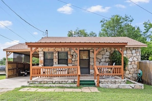 A stone house with a porch, a covered patio, and a fenced yard. - The Lazy Spur | Cozy 2BR Cabin in Cowboy Capital (Bandera)