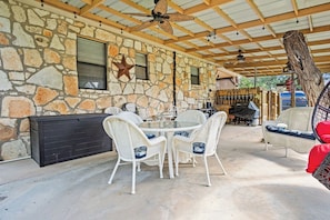 A stone-walled patio with a round table and white wicker chairs, a ceiling fan, and a wooden ceiling. - The Lazy Spur | Cozy 2BR Cabin in Cowboy Capital (Bandera)