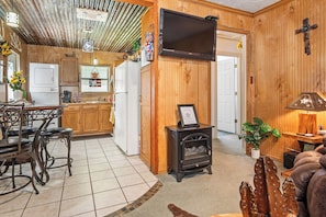 A cozy interior with a wooden dining area, a black wood-burning stove, and a television mounted on the wall. - The Lazy Spur | Cozy 2BR Cabin in Cowboy Capital (Bandera)