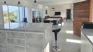 A modern kitchen with a large island, a dining area with a table and chairs, and a view of the outdoors through large windows. - "PARADISE". (Coronel Moldes)