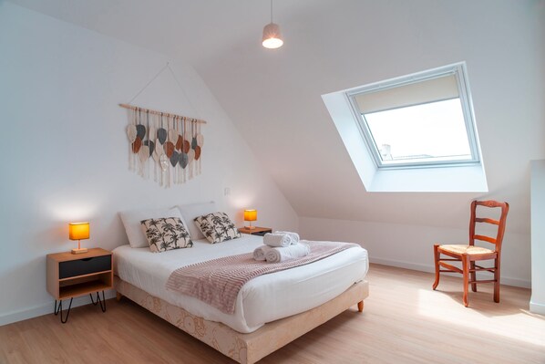 Un dormitorio con techo inclinado, tragaluces, cama con ropa de cama blanca, dos mesitas de noche con lámparas, una silla de madera y un adorno en la pared.