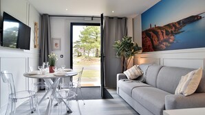 A modern living room with a grey sofa, clear chairs, a dining table with a potted plant, and a large painting of a coastal scene. - Lya 04 - 5min à Pied de la Plage (Fréhel)