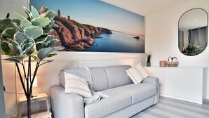 A living room with a grey sofa, a small side table, a potted plant, a wall-mounted mirror, and a large painting of a coastal landscape. - Lya 04 - 5min à Pied de la Plage (Fréhel)