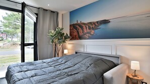 A bedroom with a large bed, a view of the outdoors through a sliding door, a painting of a coastal scene, and a bedside table with a lamp. - Lya 04 - 5min à Pied de la Plage (Fréhel)