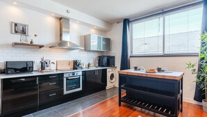 Una cocina moderna con armarios negros, microondas, estufa y lavadora. Hay una ventana con persianas y una planta a la derecha.