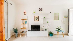 Una sala de estar moderna con un sofá blanco, televisión y estanterías. Hay plantas, una escalera y una mesita.