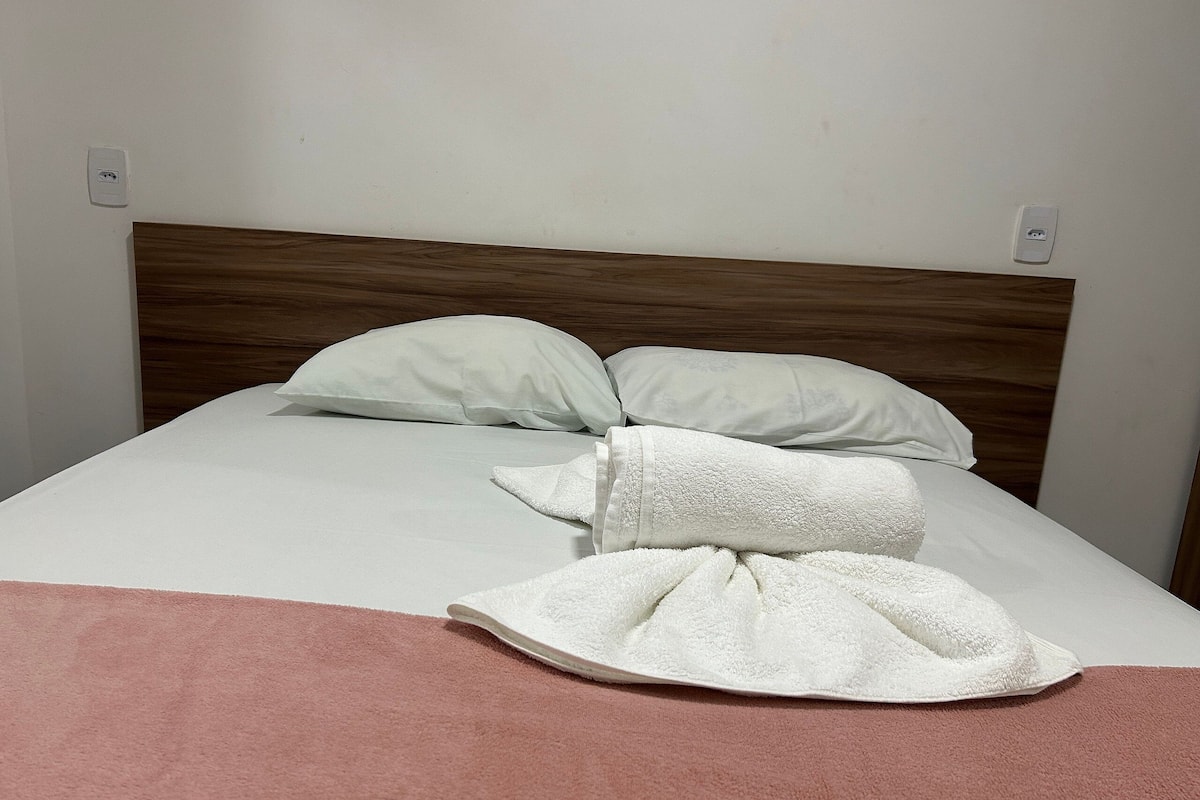 A neatly made bed with white linens and a pink blanket, a wooden headboard, and two wall sockets.