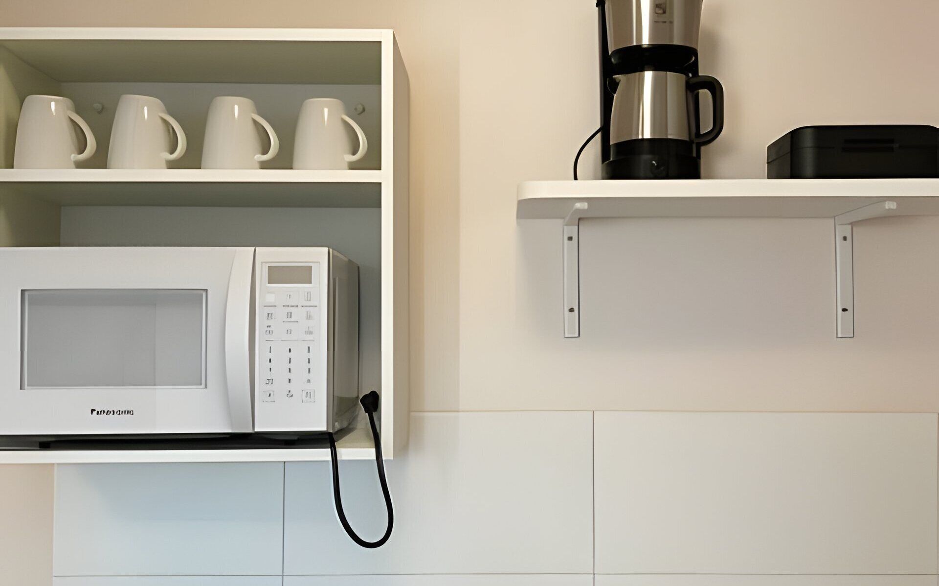 A kitchen with a white microwave, a coffee maker, and a shelf with four white cups.