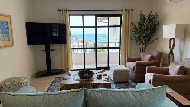 A living room with a sofa, chairs, a television, and a view of the sea through large windows. - Oceanview Apt, 3BR/2BA in The Catalinas, 5 min walk to the beach, free parking (Las Catalinas)