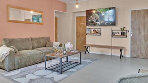 A living room with a sofa, coffee table, and TV. There are framed pictures on the wall and a door in the background. - Gîtes de France® - Green Station (FORGES LES EAUX)