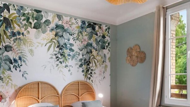 A bedroom with a bed, two wooden headboards, a floral wall mural, a window with curtains, and a radiator. - Gîtes de France® - Green Station (FORGES LES EAUX)